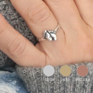 May include: A silver ring with a design of two intertwined baby feet. The ring is shown on a person's finger. The ring is also available in gold and rose gold.