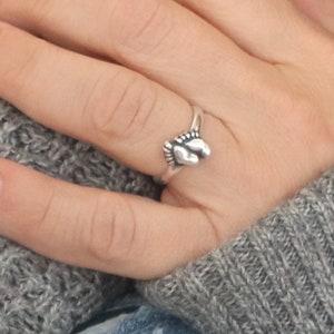 May include: A silver ring with a small footprint design. The ring is worn on a person's finger.