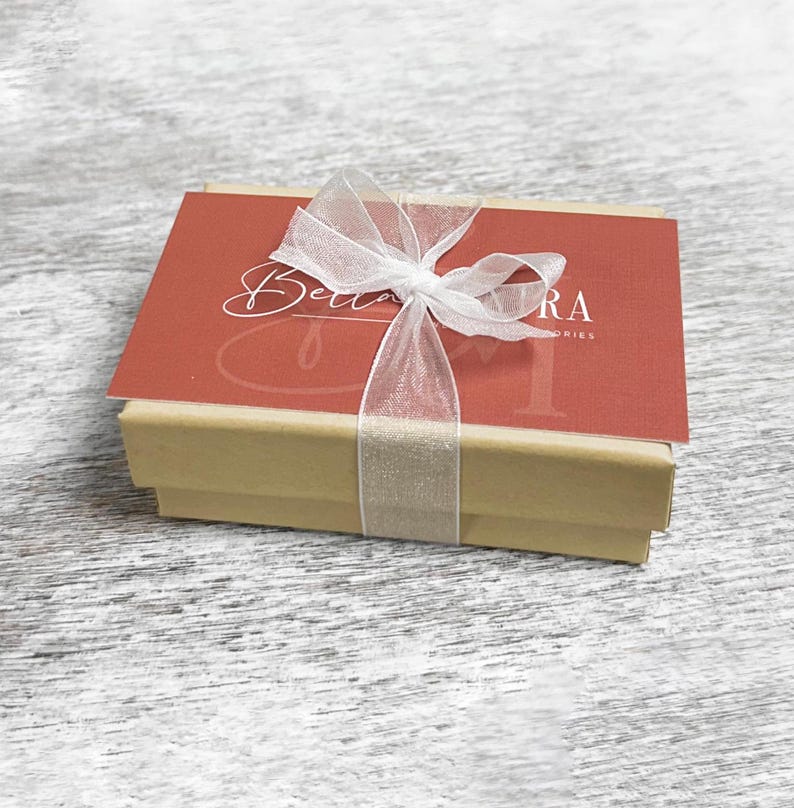 May include: A small, brown gift box with a white ribbon and bow. The box is tied with a sheer white ribbon and has a red card on top with the text "Bella Flora" in white cursive.