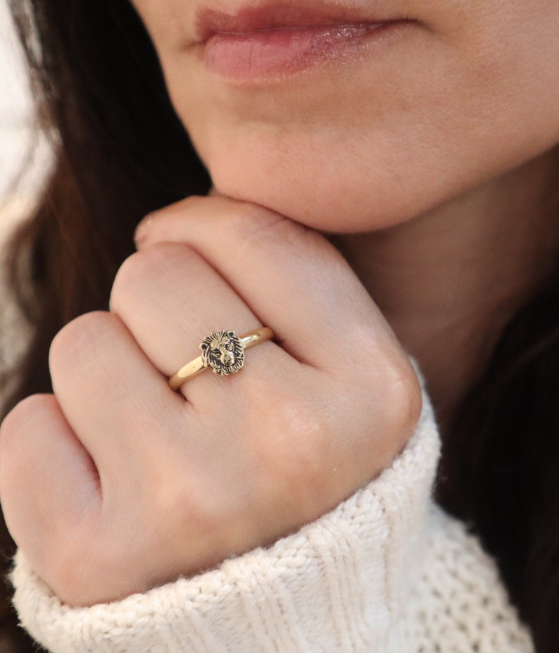 May include: A gold ring with a lion head design. The ring is worn on a person's finger.