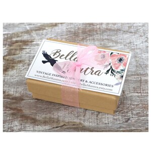 Op de afbeelding: Een kleine, rechthoekige, bruine geschenkdoos, vastgebonden met een doorschijnend roze lint. De doos heeft een wit label met de tekst "Bella Mantra" en bloemmotieven. De doos staat op een lichtbruin, houten oppervlak.