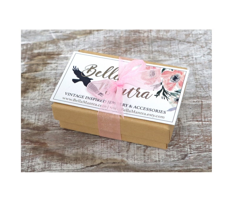 Op de afbeelding: Een kleine, rechthoekige, bruine geschenkdoos, vastgebonden met een doorschijnend roze lint. De doos heeft een wit label met de tekst "Bella Mantra" en bloemmotieven. De doos staat op een lichtbruin, houten oppervlak.