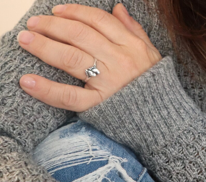 May include: A silver ring with a small footprint design. The ring is worn on a person's finger.