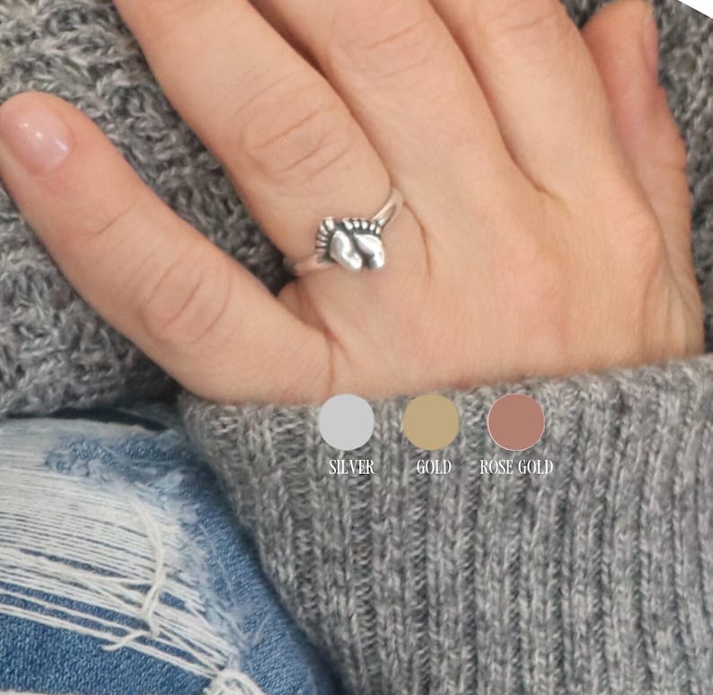 May include: A silver ring with a design of two intertwined baby feet. The ring is shown on a person's finger. The ring is also available in gold and rose gold.