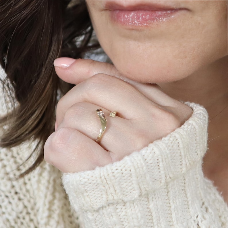 May include: A gold ring with a textured band and a small square accent. The ring is open and can be adjusted to fit different finger sizes.