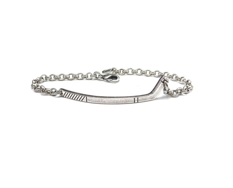 Op de afbeelding: Een zilveren armband met een staafvormig middelpunt en een schakelketting. De staaf heeft ge&euml;tste lijnen en rechthoekige details. De armband wordt op een witte achtergrond getoond.