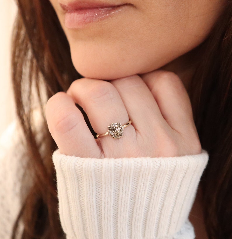 May include: A gold ring with a lion head design. The ring is worn on a person's finger.