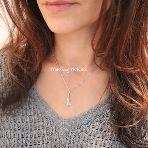 May include: A silver necklace with a small silver cello pendant. The necklace is on a model wearing a gray sweater. The text "Matching Necklace" is visible on the image.