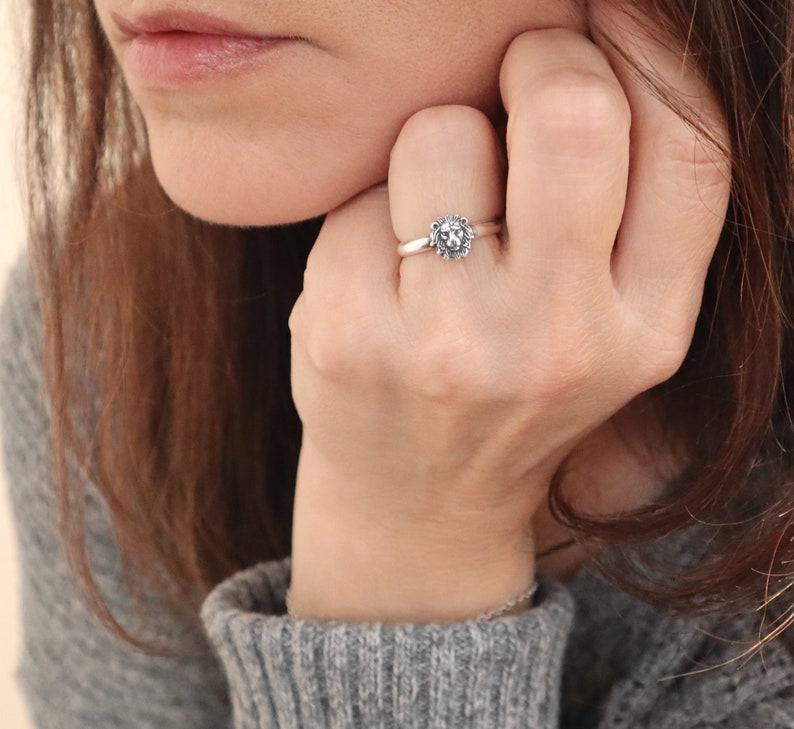 May include: A silver ring with a lion head design. The ring is worn on a person's finger.