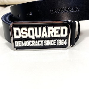 May include: Black leather belt with a silver buckle. The buckle features the brand name "DSQUARED" in bold white letters, with "DEMOCRACY SINCE 1964" below. The belt is on a white surface.