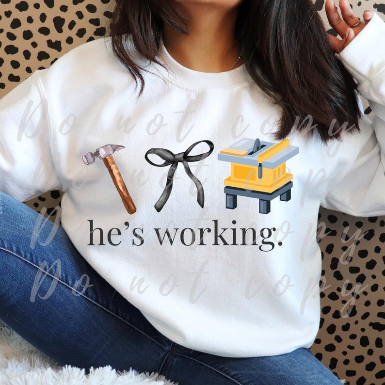 May include: White sweatshirt with a hammer, bow, and table saw graphic. The text "he's working" is printed below the graphics. The sweatshirt is a casual, everyday garment.