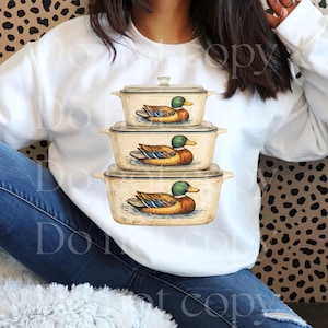 May include: White sweatshirt featuring a stack of three vintage-style casserole dishes. Each dish has a duck illustration. The ducks have green heads, brown bodies, and blue and yellow accents. The dishes have glass lids.
