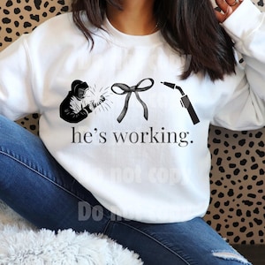May include: White sweatshirt with a black graphic of a welder, a bow, and a welding torch. The text "he's working" is printed below the graphics. The sweatshirt is worn by a person wearing blue jeans.