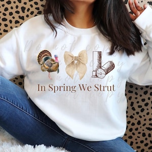Könnte beinhalten: Weißer Pullover mit einem braunen Truthahn, einer braunen Schleife und braunen Schrotpatronen. Der Text "In Spring We Strut" ist auf dem Pullover aufgedruckt.
