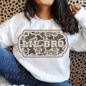 May include: White sweatshirt with a camouflage design and the text "LIL BRO" in a distressed font. The graphic is in a rectangular shape with rounded corners. The sweatshirt is a casual style.