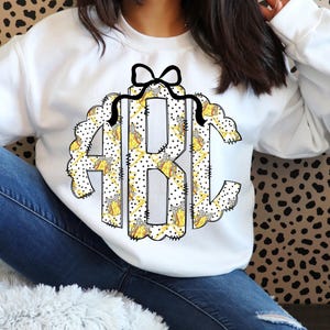 May include: White sweatshirt with a large monogram design featuring the letters "ABC". The monogram is framed by a scalloped edge and a black bow. The letters are filled with a yellow and white pattern with black polka dots.