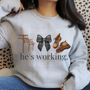 May include: A light gray sweatshirt with a graphic design. The design includes brown utility poles, a black bow, and brown boots. The text "he's working" is printed below the graphics.