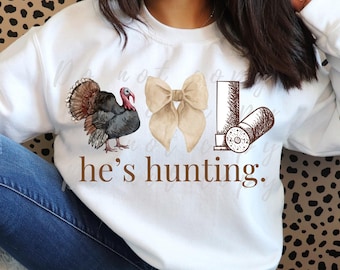 May include: White sweatshirt with a brown turkey, a brown bow, and a brown shotgun shell graphic. The text "he's hunting." is printed below the graphics.