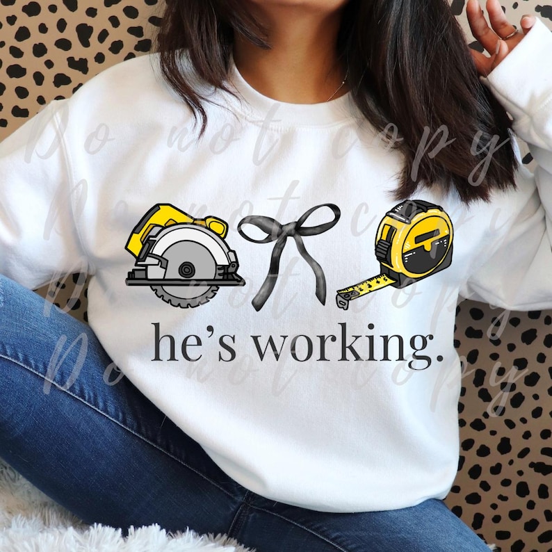 May include: White sweatshirt with a graphic of a circular saw, a bow, and a tape measure. The text "he's working" is printed below the graphics. The saw is yellow and gray, the bow is black, and the tape measure is yellow and black.