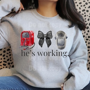 May include: A light gray sweatshirt featuring a red welding machine, a black bow, and a welding helmet. The text "he's working" is printed below the graphics. The design is a watercolor style.