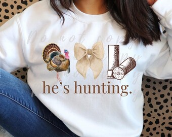 May include: White sweatshirt with a brown turkey, a brown bow, and a brown shotgun shell with the text "he's hunting."
