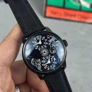 May include: A black Jacob & Co. wristwatch with a black fabric strap. The watch face has multiple dials with white numerals and a world map design. The watch is held against a blurred background.