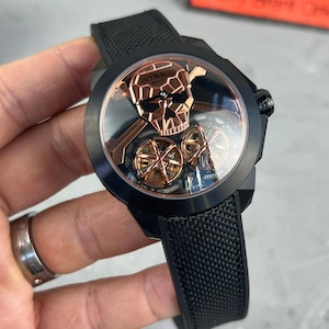 May include: Black wristwatch with a skull design and rose gold detailing. The watch has a black strap and a transparent face, revealing the internal mechanisms. The watch face displays the brand name "JACOB & CO."