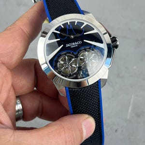 以下が含まれることがあります： 黒い文字盤とシルバーのベゼルを備えた高級腕時計。 時計には「JACOB & CO GENEVE」のブランド名と黒と青のストラップが付いています。 時計には2つの内部メカニズムが見え、文字盤の周りに青いアクセントがあります。