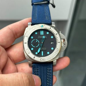 Op de afbeelding: Een Panerai Submersible horloge met een matte zilverkleurige kast, een zwarte wijzerplaat met lichtblauwe lichtgevende uurmarkeringen, een datumvenster op de drie-uurpositie en een gestructureerde blauwe stoffen band met gespsluiting.