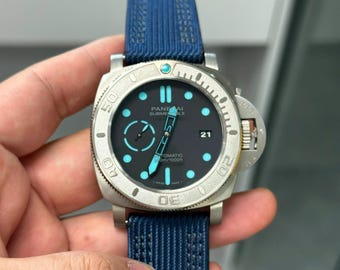 May include: A Panerai Submersible watch with a matte silver-tone case, a black dial featuring luminous light blue hour markers, a date window at the three o'clock position, and a textured blue fabric strap with a secure buckle closure system.