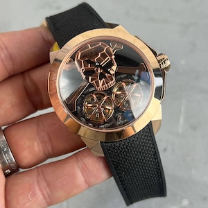 May include: Rose gold wristwatch with a black strap. The watch face features a skull design and visible internal mechanisms. The watch has a round face and a textured black band.
