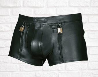 Könnte beinhalten: Schwarze Shorts in Lederoptik mit strukturiertem Bund, zwei Druckknöpfen, einem konturierten Mittelteil und zwei vertikalen Reißverschlusstaschen auf der Vorderseite. Jeder Reißverschlussanhänger ist mit einem kleinen, rechteckigen Metallschloss versehen.