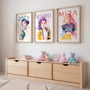 May include: Three framed watercolor prints with the names "ZOEY", "RUMI", and "MIRA" above stylized portraits of women. A light wood dresser with three drawers sits below the prints, with stuffed animals and a lollipop on top.
