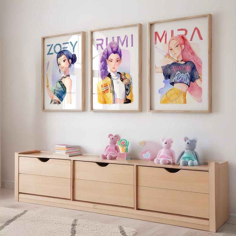 May include: Three framed watercolor prints with the names "ZOEY", "RUMI", and "MIRA" above stylized portraits of women. A light wood dresser with three drawers sits below the prints, with stuffed animals and a lollipop on top.