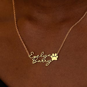 Puede incluir: Un collar de tono dorado con una cadena fina y un colgante personalizado. El colgante muestra los nombres Evelyn y Barry escritos en una fuente cursiva, acompañados por un pequeño icono de huella de animal sólido situado a la derecha.