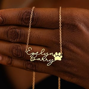 Puede incluir: Un collar dorado con el nombre "Evelyn Baby" en cursiva y un dije de huella de pata. El collar lo sostiene una persona de piel oscura. La cadena es delicada y el colgante es un diseño personalizado.