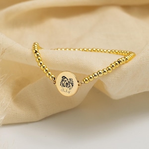 May include: Gold beaded bracelet with a circular charm. The charm features a black illustration and the word "July" in script. The bracelet is displayed on a cream-colored fabric.