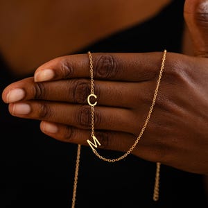 Puede incluir: Un collar dorado con las letras "C" y "M". El colgante cuelga de una fina cadena, creando un accesorio personalizado y elegante. El collar se muestra sobre un fondo oscuro, resaltando el color dorado.