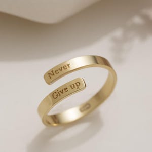 Puede incluir: Un anillo dorado con las palabras "Never Give up" grabadas en la banda. El anillo tiene un diseño abierto, con los extremos ligeramente superpuestos. El anillo se muestra sobre una superficie blanca con flores secas en el fondo.
