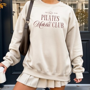 Könnte beinhalten: Beiges Sweatshirt mit burgunderfarbenem Schriftzug "PILATES Matcha CLUB". Dazu ein passender Faltenrock und weiße kniehohe Stiefel. Eine braune Umhängetasche und ein weißer Kaffeebecher ergänzen das Outfit.