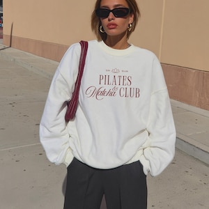 Puede incluir: Sudadera color crema con el texto "PILATES & Matcha CLUB" en escritura burdeos. La sudadera tiene cuello redondo y mangas largas. La persona lleva gafas de sol negras y pantalones negros.