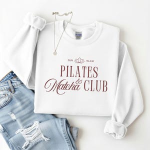 May include: A white crewneck sweatshirt with the words "PILATES Matcha & CLUB" in burgundy script. The sweatshirt is paired with light wash denim jeans and a silver necklace.
