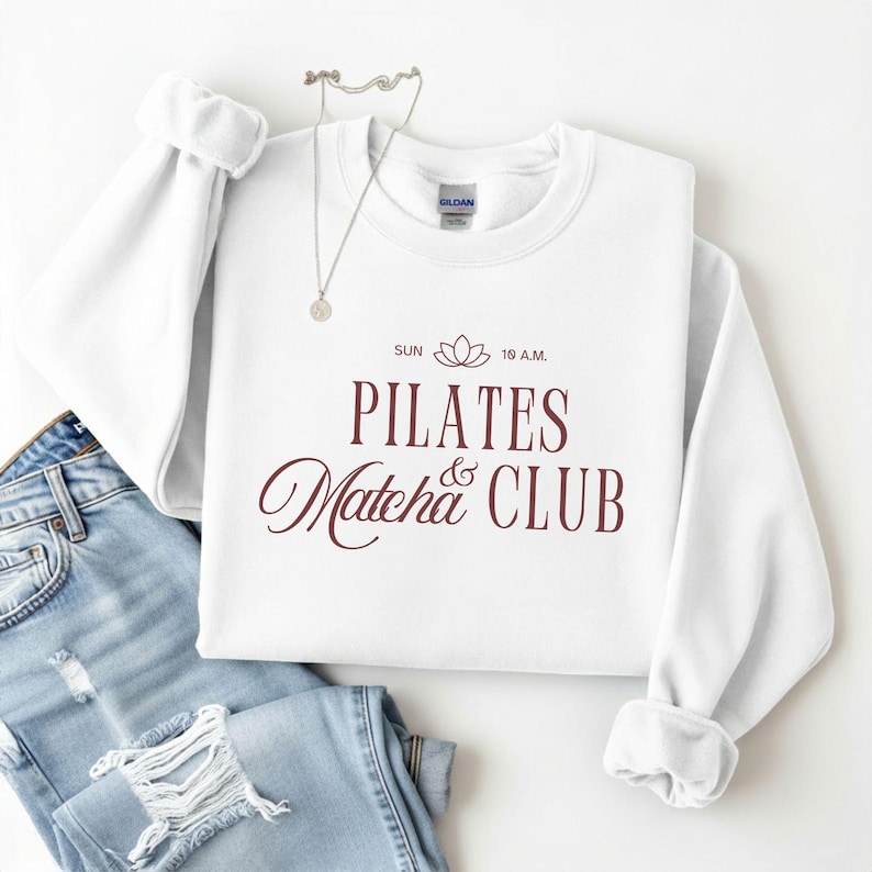 May include: A white crewneck sweatshirt with the words "PILATES Matcha & CLUB" in burgundy script. The sweatshirt is paired with light wash denim jeans and a silver necklace.