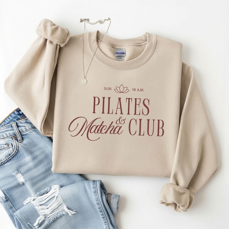 May include: A beige sweatshirt with the words "PILATES Matcha CLUB" in burgundy script. The text includes a lotus flower graphic and the time "SUN 10 A.M." The sweatshirt is folded with a pair of blue jeans.
