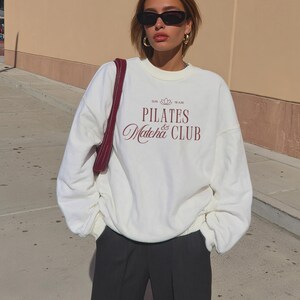 May include: A cream-colored sweatshirt with the text "PILATES & Matcha CLUB" in burgundy script. The sweatshirt is paired with black pants. The image also shows a burgundy shoulder bag, black sunglasses, and gold hoop earrings.