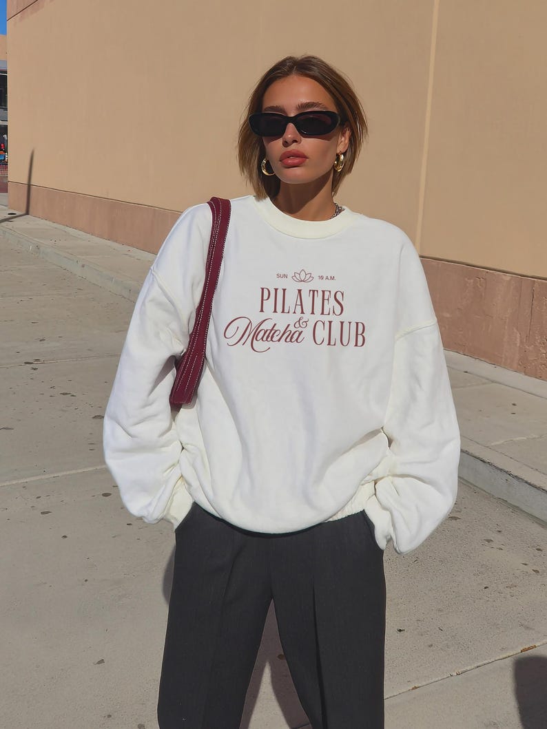 May include: A cream-colored sweatshirt with the text "PILATES & Matcha CLUB" in burgundy script. The sweatshirt is paired with black pants. The image also shows a burgundy shoulder bag, black sunglasses, and gold hoop earrings.