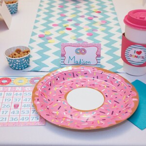 May include: A pink and white donut-themed party table setting with a pink and white donut plate, a pink and white donut bingo card, a pink and white donut cup, a pink and white donut napkin, and a pink and white donut name card that says "Madison".