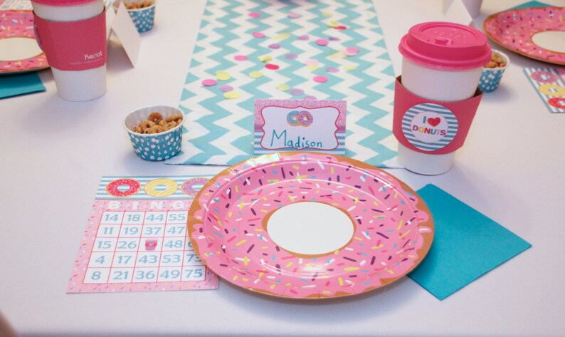 May include: A pink and white donut-themed party table setting with a pink and white donut plate, a pink and white donut bingo card, a pink and white donut cup, a pink and white donut napkin, and a pink and white donut name card that says "Madison".