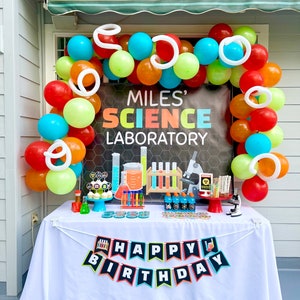 May include: A birthday party table decorated with a science theme. The table is covered with a white tablecloth and has a banner that says "Happy Birthday". There are balloons in various colors, including red, orange, yellow, green, blue, and white. The table is decorated with science-themed decorations, such as beakers, test tubes, and a microscope. The backdrop is a black and white banner that says "Miles' Science Laboratory".