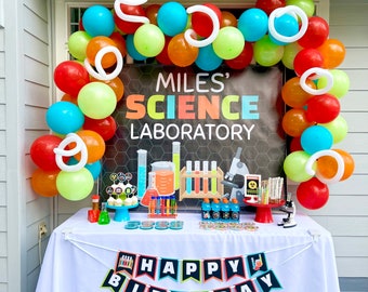 May include: A birthday party table decorated with a science theme. The table is covered with a white tablecloth and has a banner that says "Happy Birthday". There are balloons in various colors, including red, orange, yellow, green, blue, and white. The table is decorated with science-themed decorations, such as beakers, test tubes, and a microscope. The backdrop is a black and white banner that says "Miles' Science Laboratory".